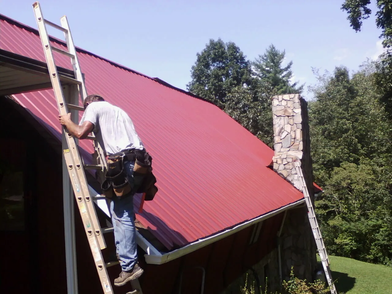 Roofer on a ladder beside a red metal roof and stone chimney — Soffit & Fascia Repair by Roofing Brothers in Pelham